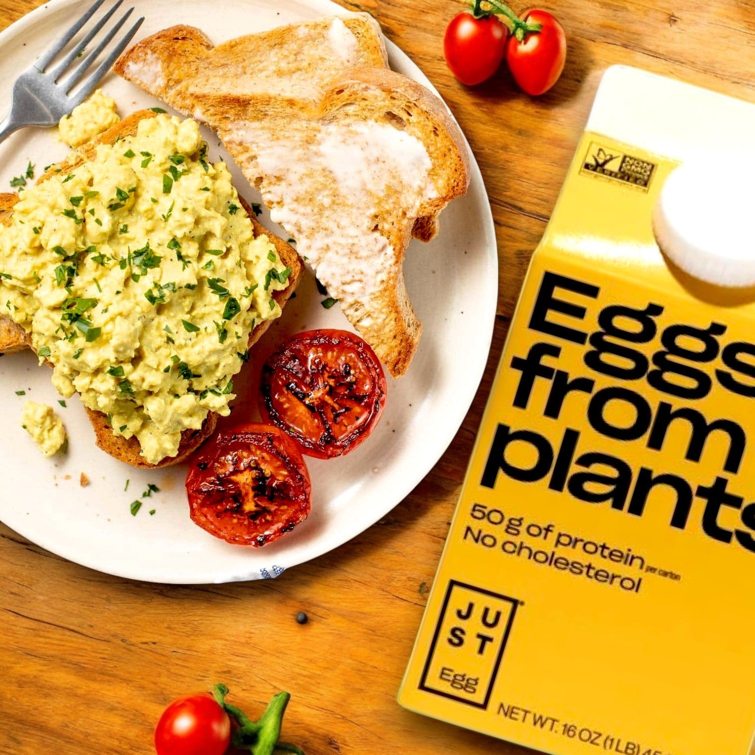 A plate features scrambled eggs on toast with roasted tomatoes and parsley, next to a box labeled "Just Egg Plant-Based Egg (16 oz)." A fork, cherry tomatoes, and parsley suggest a Just Egg meal—a cholesterol-free plant-based alternative to traditional breakfast favorites.