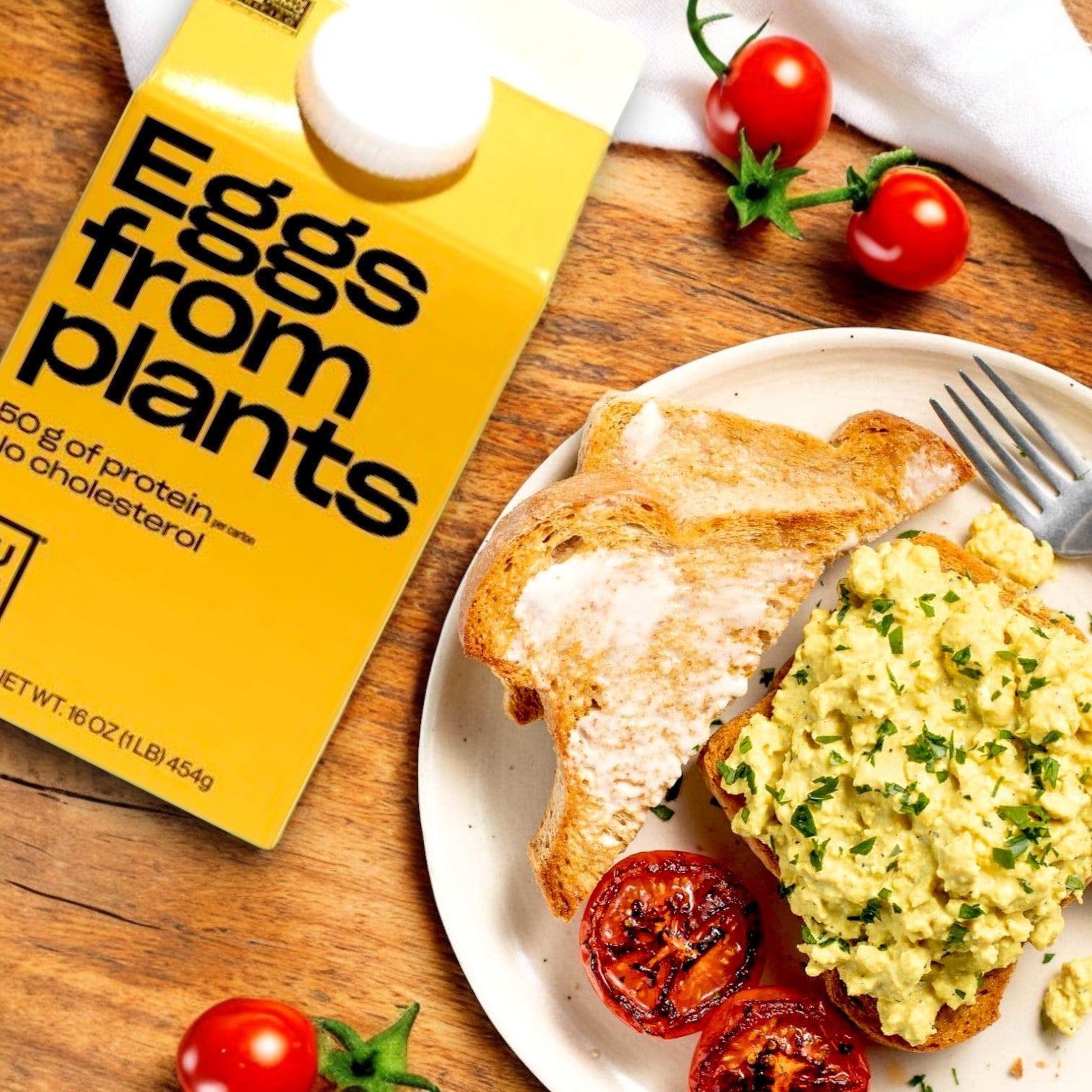 A plate with plant-based scrambled eggs on toast, roasted tomatoes, and a carton labeled "Just Egg Plant-Based Egg (16 oz)" by Just Egg sits on a wooden table scattered with cherry tomatoes. This cholesterol-free delight offers a delicious plant-based alternative for breakfast.