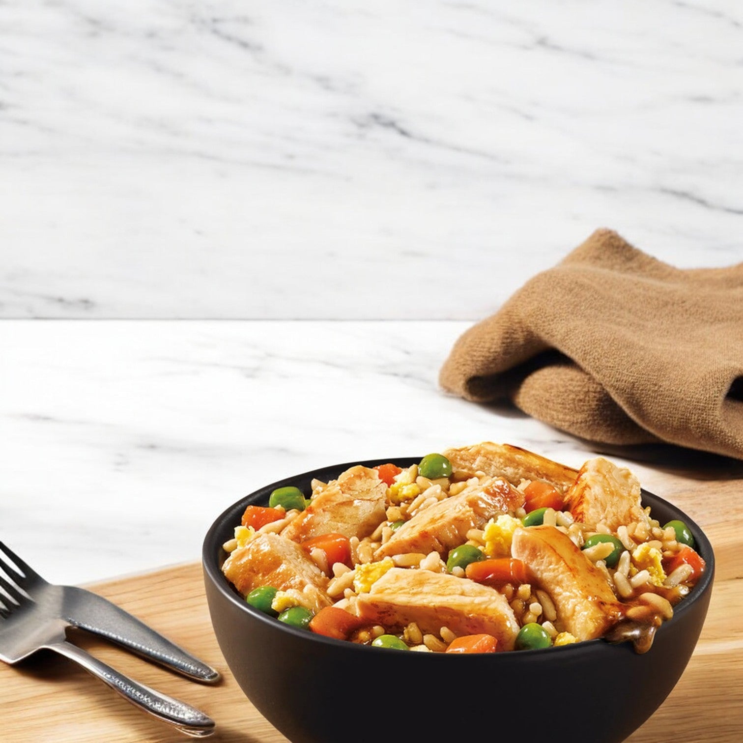 Marie Callender's Savory Chicken Fried Rice Bowl includes a delightful mix of rice, peas, carrots, corn, and chicken slices. Paired with two forks on a wooden surface, the scene is completed with a brown napkin for a homey touch—perfect for enjoying this Smart Ones frozen meal.