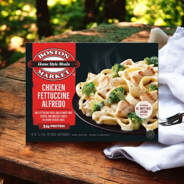 A 13 oz. box of Smart Ones' Boston Market Chicken Fettuccine Alfredo is displayed on a wooden table with a white cloth and fork, featuring pasta with chicken and broccoli in creamy Alfredo sauce for a comforting taste experience.