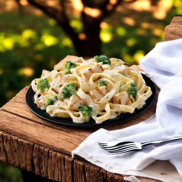 Boston Market Chicken Fettuccine Alfredo by Smart Ones, 13 oz, sits on a rustic wooden table with broccoli and a soy-based sauce. A fork on a napkin completes the outdoor setting.