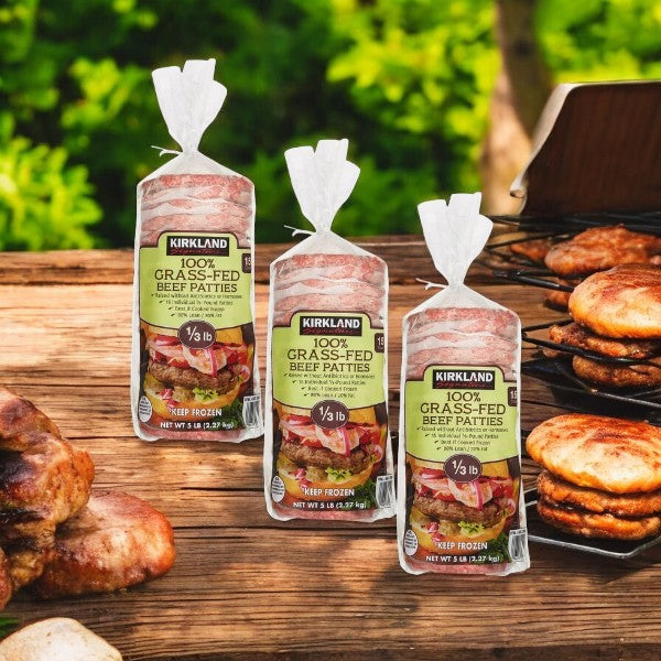 Three packs of Kirkland Signature Grass-Fed Beef Patties, each at 1/3 lb, are displayed on a wooden table outdoors with cooked patties and greenery in the background, highlighting a protein-rich meal.