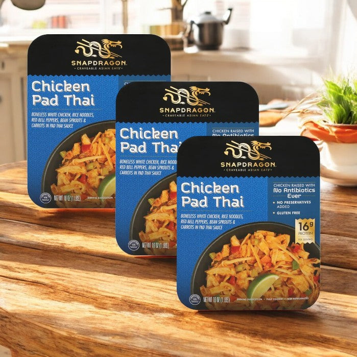 Three 32 oz packages of Snapdragon Chicken Pad Thai, each featuring a close-up image of the savory dish, are neatly displayed on a wooden table. This convenient frozen meal captures the essence of authentic Thai cuisine.
