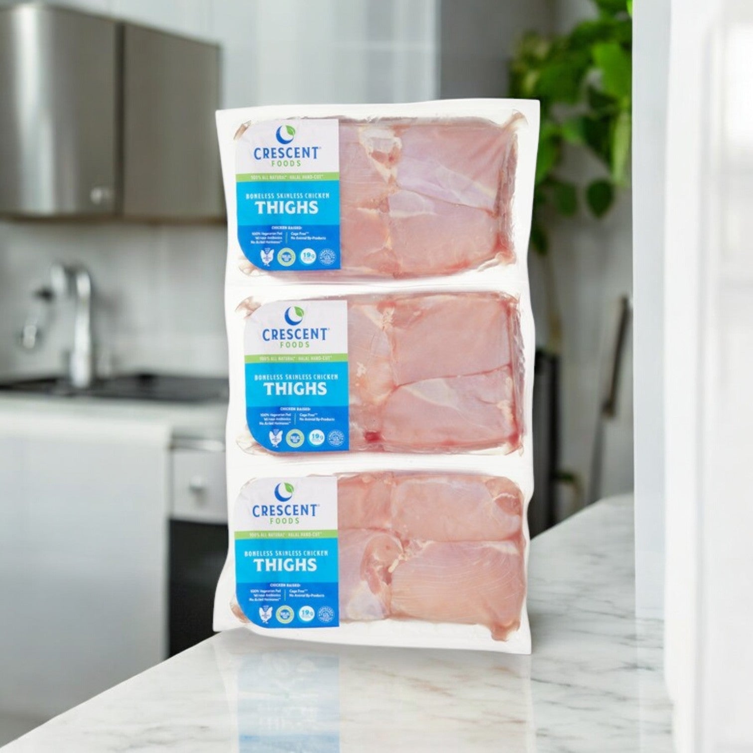 Three packages of hand-cut Crescent Foods Halal Chicken Thighs, boneless & skinless (approx. 4 lbs), are stacked on a kitchen counter with a stove and cabinets in the background.