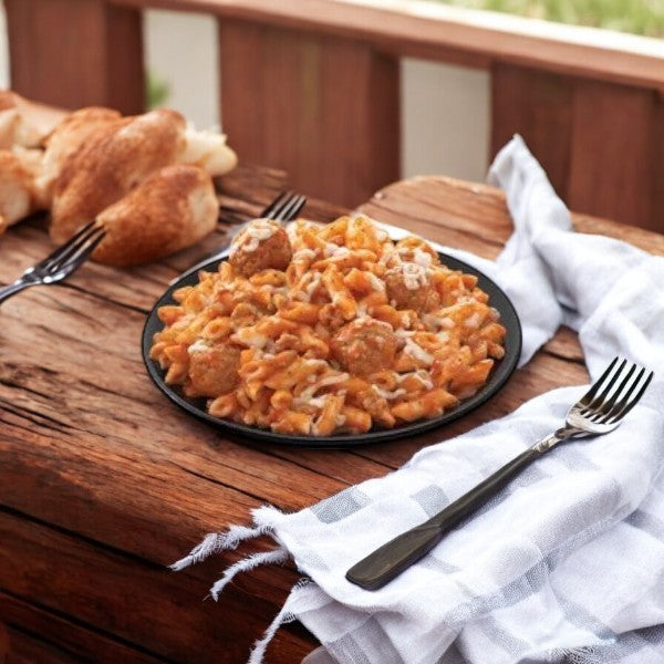 A plate of Smart Ones Boston Market Penne with Meatballs 13 oz sits on a wooden table with two forks and a blue-and-white napkin. Fresh bread in the background adds to this delightful scene.