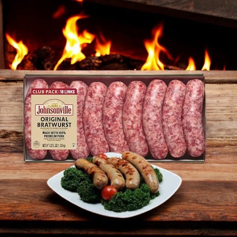 A case of Johnsonville Brats Original Pork Bratwurst Links entices by the fireplace, with grilled sausages, kale, and cherry tomatoes in front—ideal for cozy cookouts or indoor grilling.