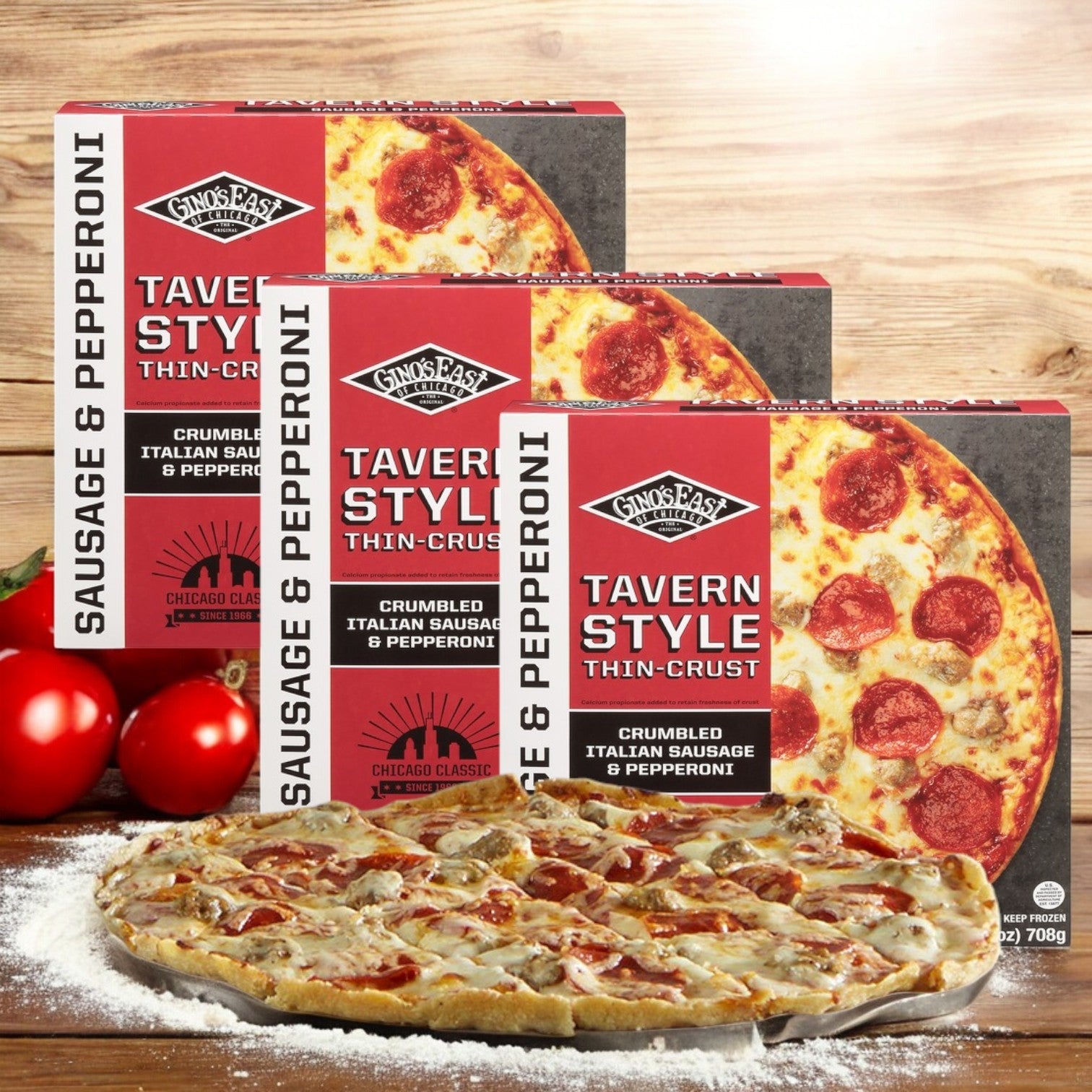Three Gino's East Thin Crust Tavern Style Sausage & Pepperoni Pizzas (25 oz - 3 Count) are lined up behind a freshly cooked pizza on a wooden surface, surrounded by ripe tomatoes and dusted with flour.