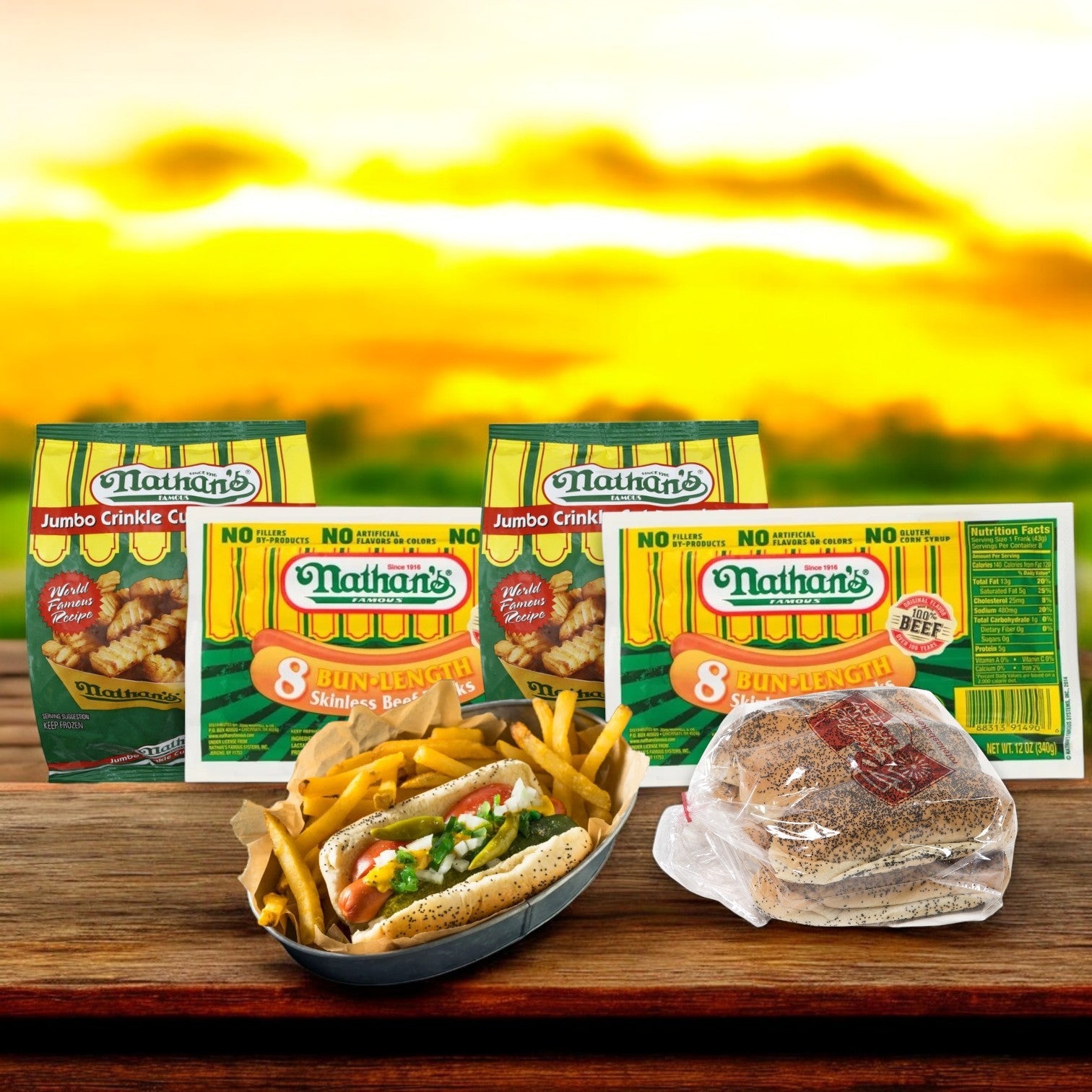 Displayed against a wooden surface with a blurred sunset backdrop is Nathan's Famous Hot Dog Bundle, featuring 16 hot dogs nestled in 16 buns alongside two bags of fries.