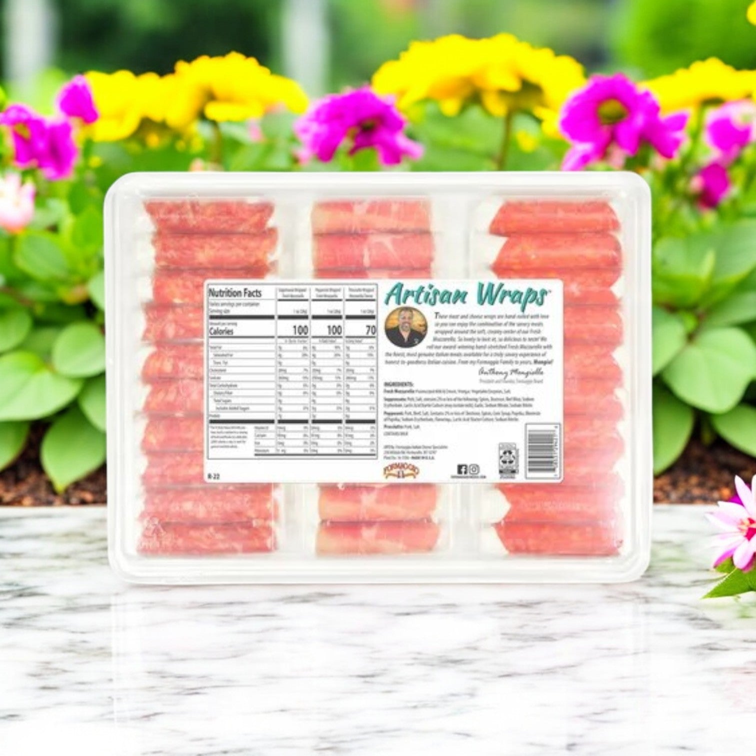 A Formaggio Artisan Wraps (22 oz) plastic package of salami-wrapped mozzarella cheese sticks sits on a marble surface, with pink and yellow flowers as a charming backdrop.