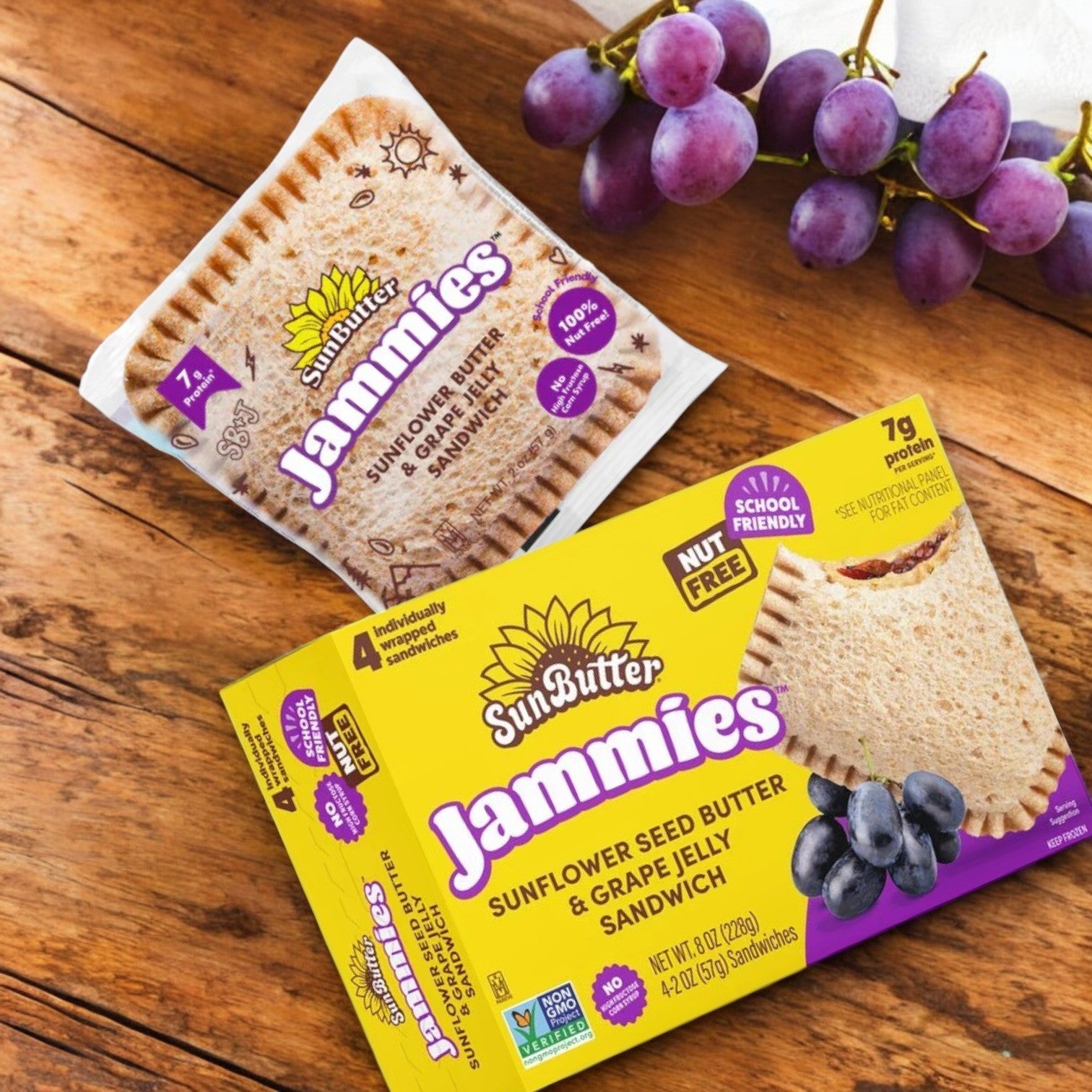 A package of SunButter Jammies 2-Pack Combo – Delicious & Nut-Free! from Sunbutter is on a wooden surface, showcasing a sunflower seed butter and grape jelly sandwich with fresh grapes.