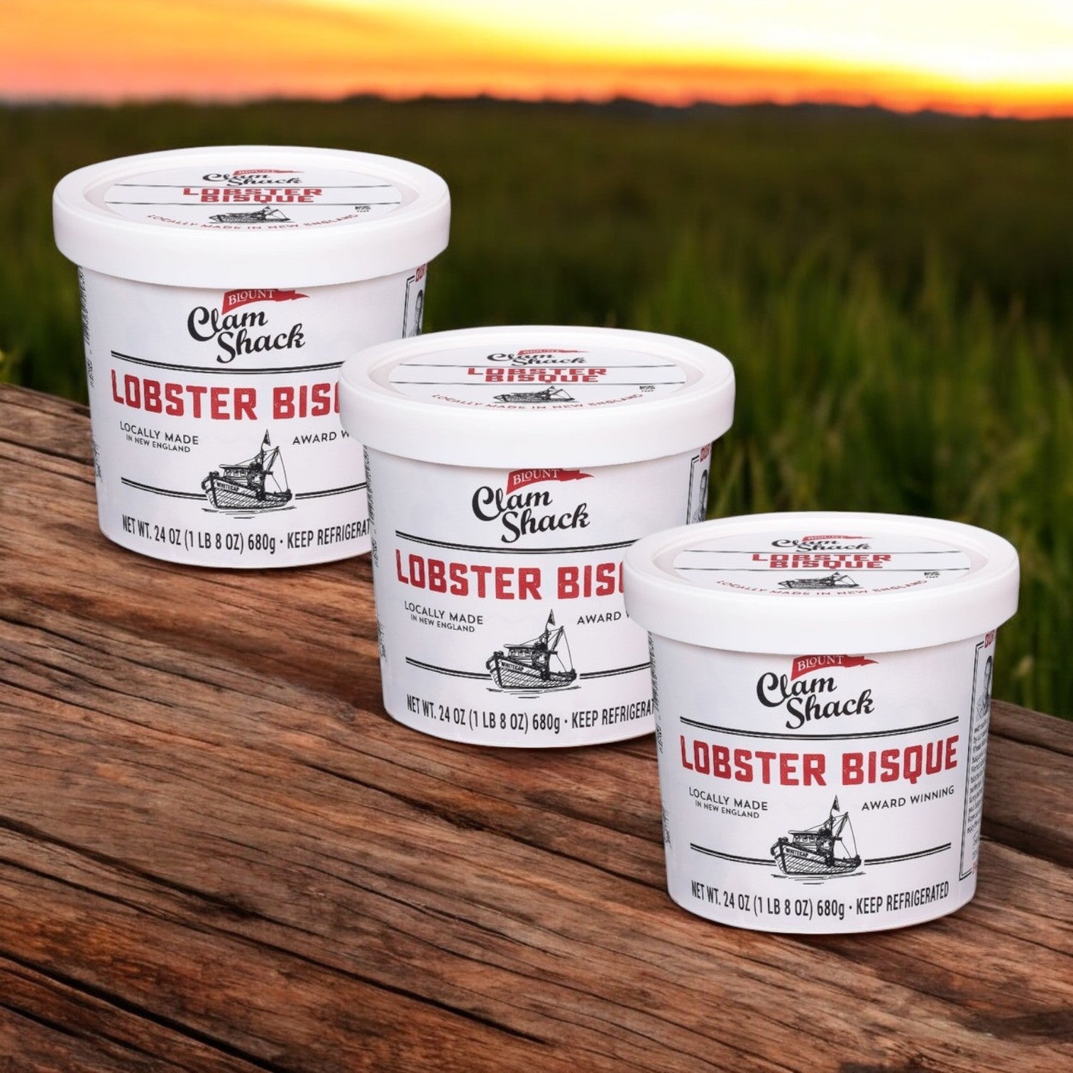 Three tubs of Blount Clam Shack Lobster Bisque Soup, a gourmet delight from Blount Fine Foods, rest on a wooden surface with a grassy field and sunset in the background.