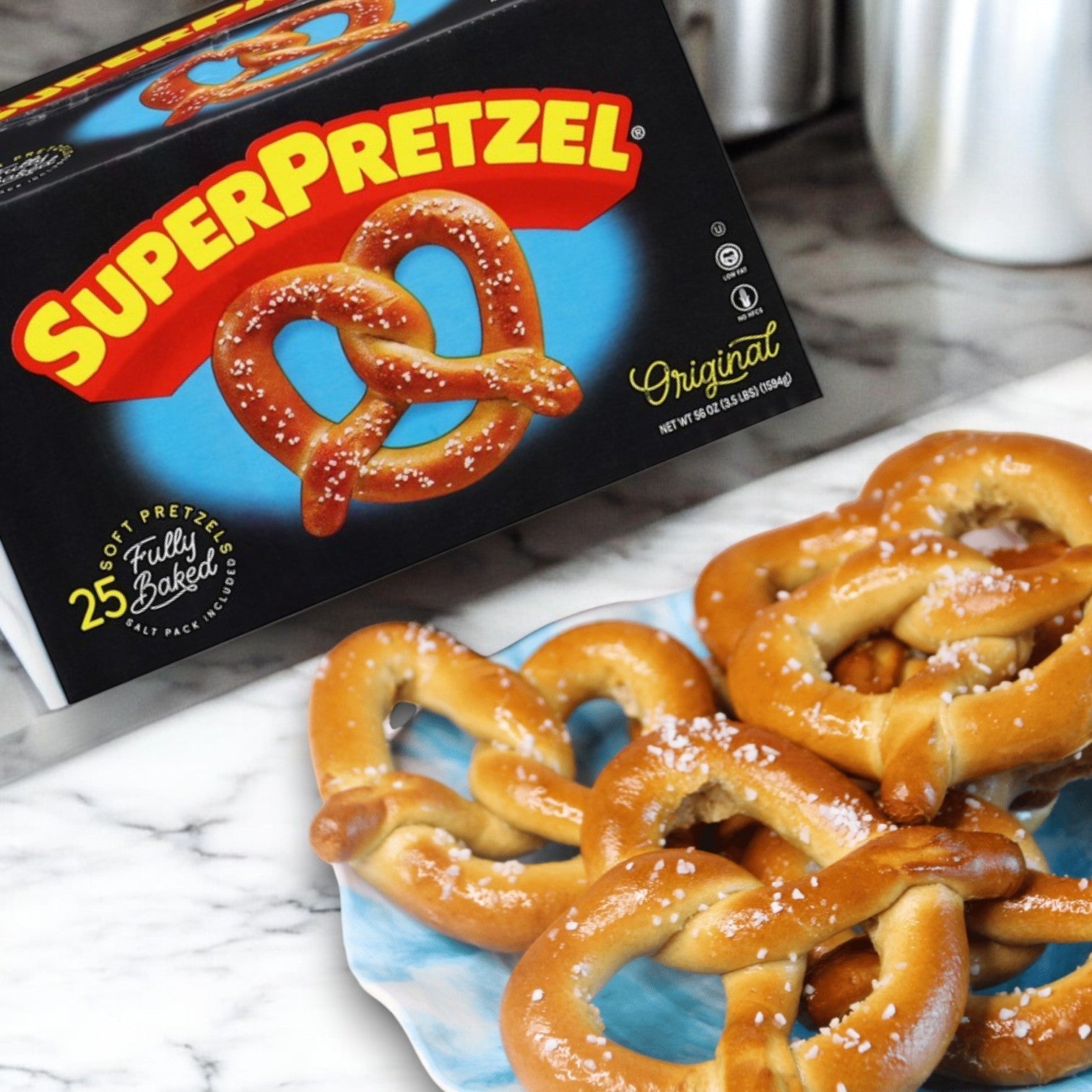 A box of SUPERPRETZEL Original Soft Pretzels (25 Count) and several salted pretzels on a blue plate are shown on a marble countertop, providing an ideal quick snack in a family-sized pack.