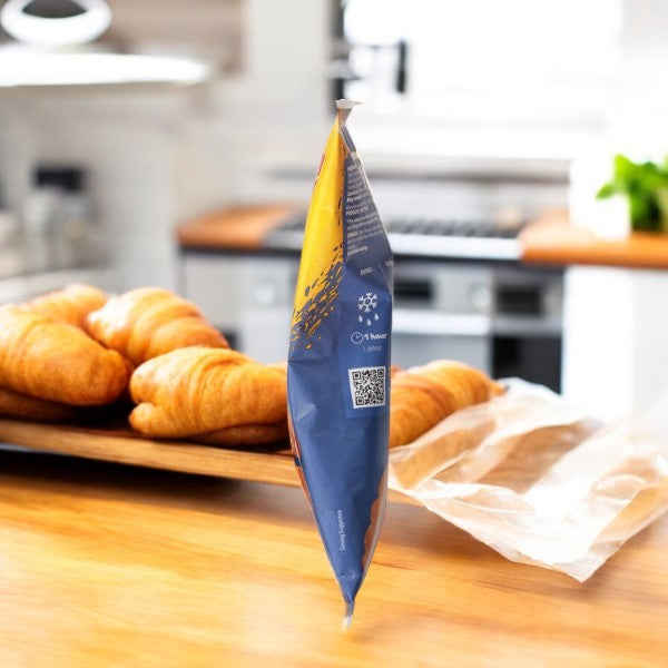 A partially empty coffee bag in shades of blue and orange stands upright on a wooden kitchen counter, surrounded by Schar Plain Croissants, 7.8 oz - 1 Per Case, which have a light and flaky texture as they rest against a blurred background.