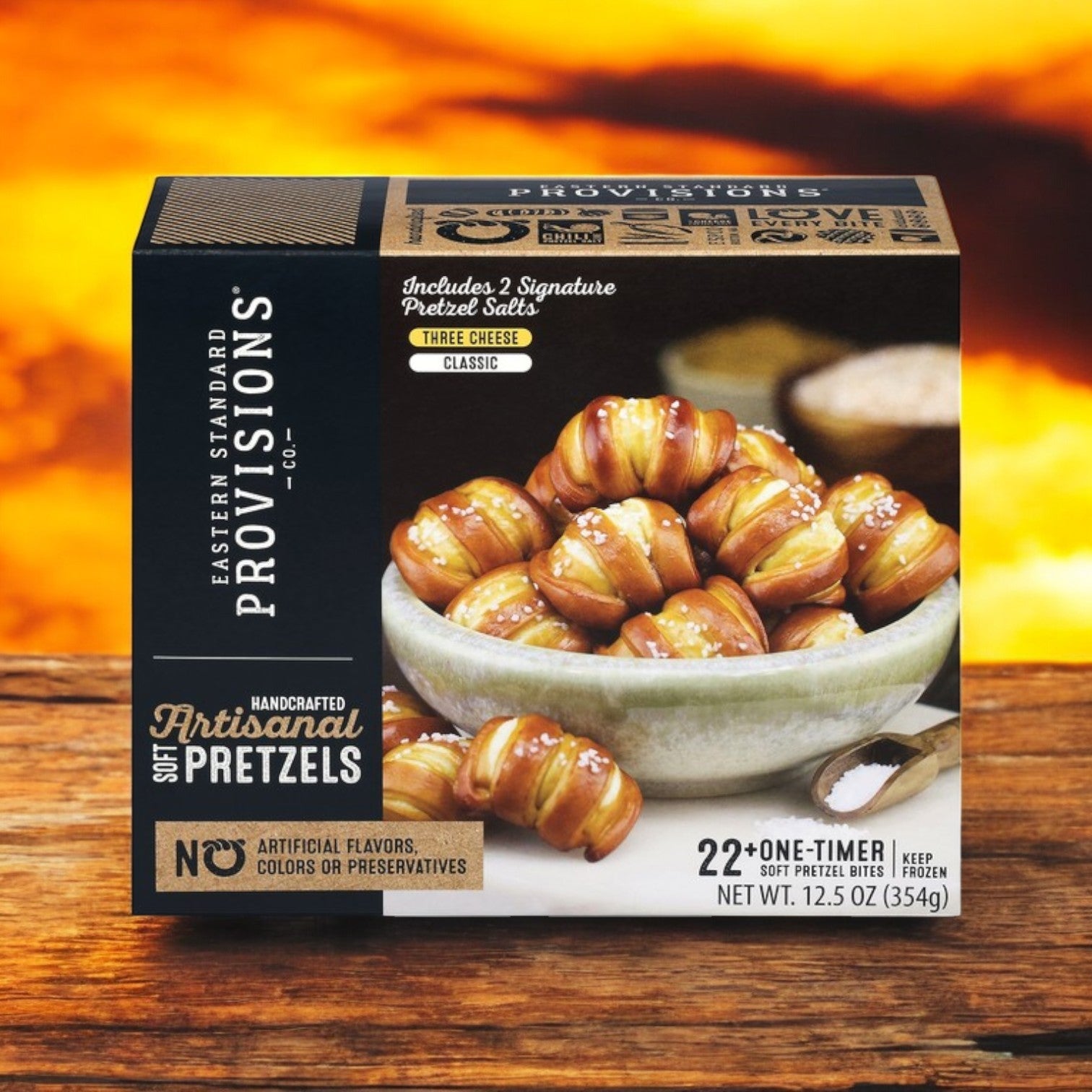 A box of Eastern Standard Provisions Pretzel 3-Pack Combo, featuring artisanal soft pretzels in three cheese and classic flavors, rests on a wooden surface with a blurred warm background; net weight is 12.5 oz (354g).