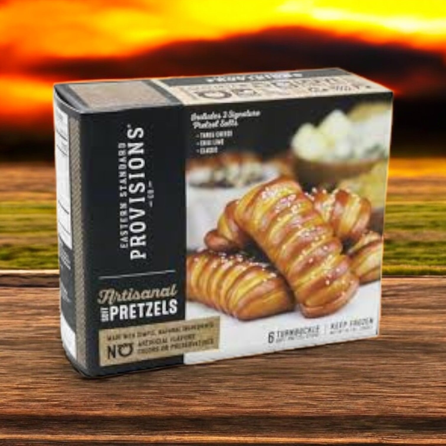 An Eastern Standard Provisions Pretzel 3-Pack Combo rests elegantly on a wooden surface against a blurred sunset backdrop, perfectly embodying the savory allure of their artisanal soft pretzels.