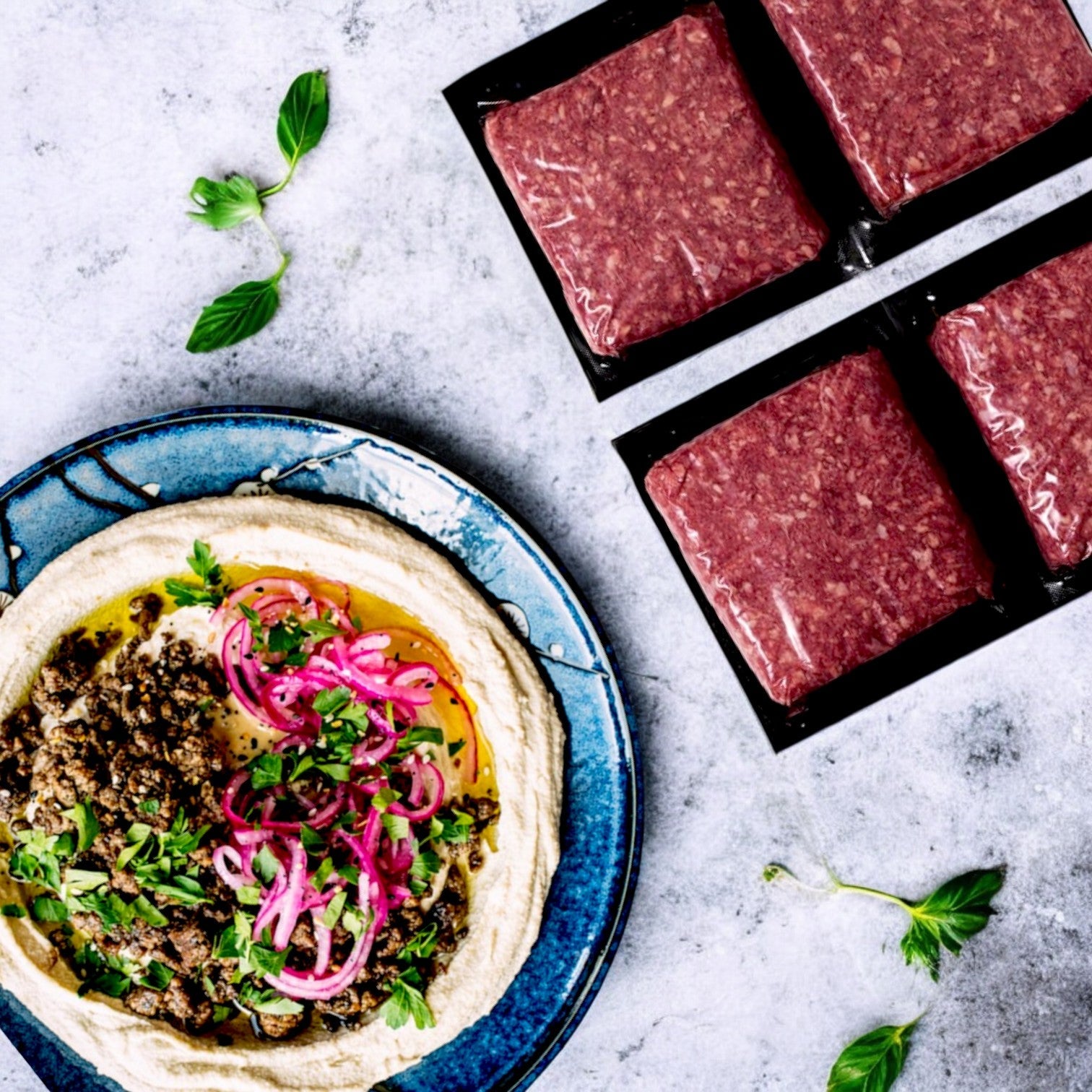 A plate with hummus, seasoned meat, and pickled onions sits beside four Diamond Valley Halal Ground Beef chubs (85% lean, 1 lb) from New Zealand Spring Lamb on a textured surface sprinkled with herbs.
