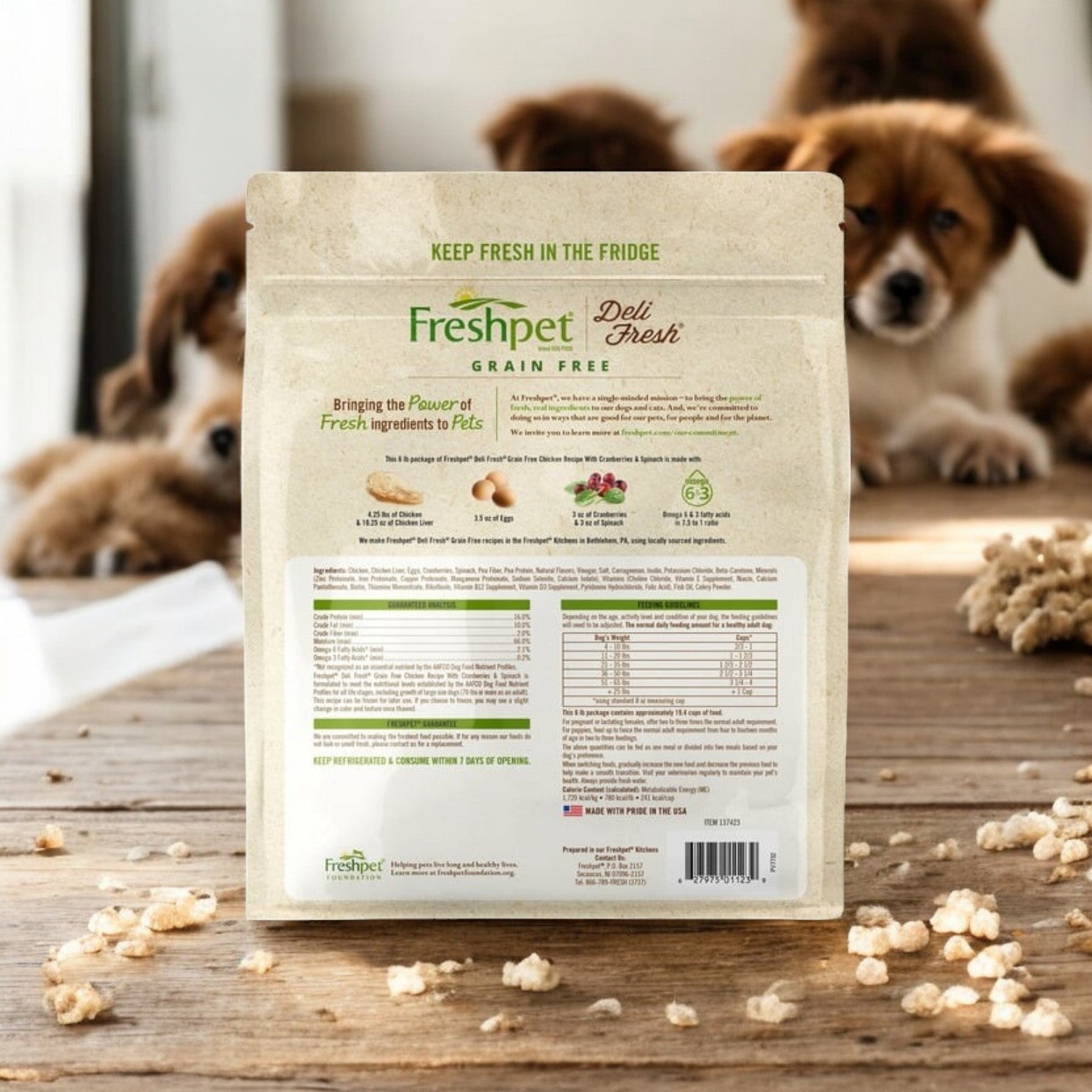 Three boxes of Freshpet Deli Fresh Dog Food, Chicken with Cranberries & Spinach, are on the table as playful puppies frolic in the background.