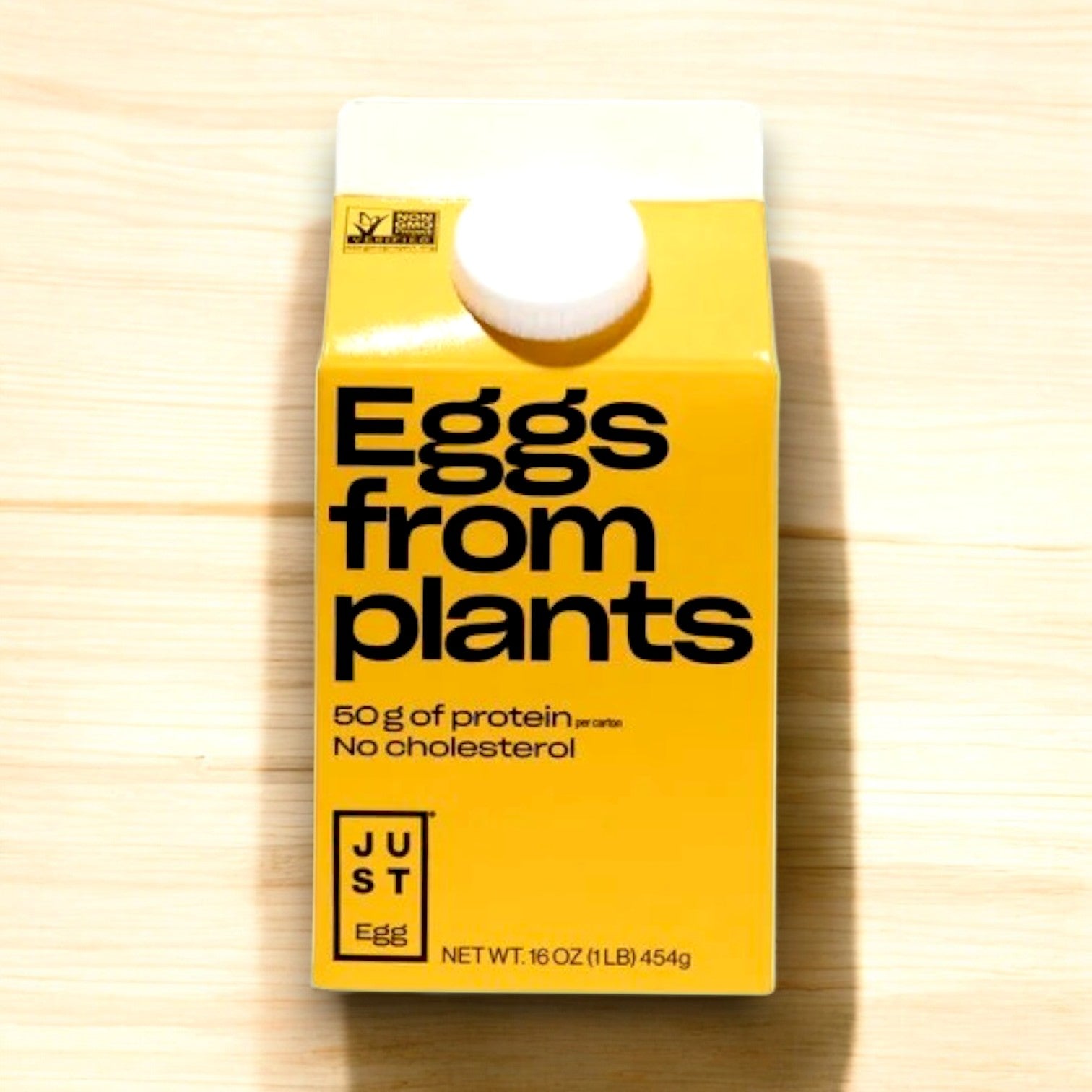 A yellow carton titled "Just Egg Plant-Based Egg (16 oz)" from Just Egg, highlights its 50g of protein and cholesterol-free, plant-based benefits, displayed on a light wooden background.