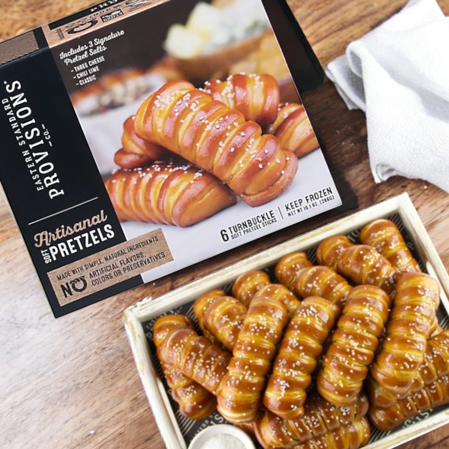 A 6-count box of Eastern Standard Provisions Soft Artisanal Turnbuckle Pretzels sits beside a tray of artisanal soft pretzels on a table adorned with a white napkin.