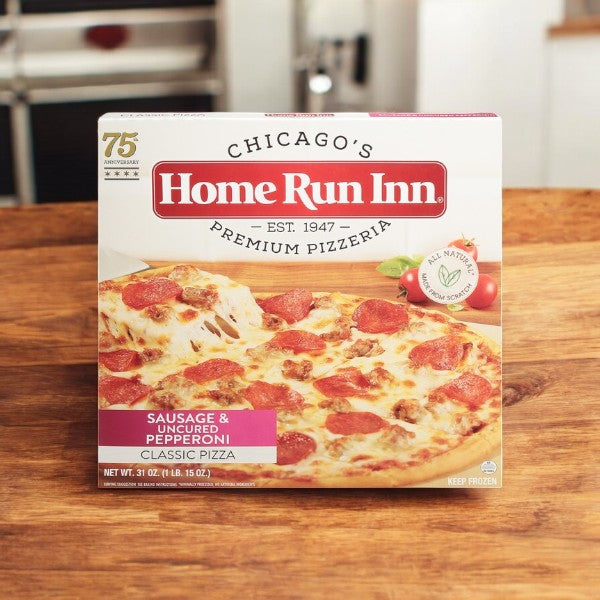 On a wooden surface lies a pizza box marked "Home Run Inn," displaying their Family Size Classic Frozen Sausage and Uncured Pepperoni Pizza. The label celebrates the 75th anniversary.