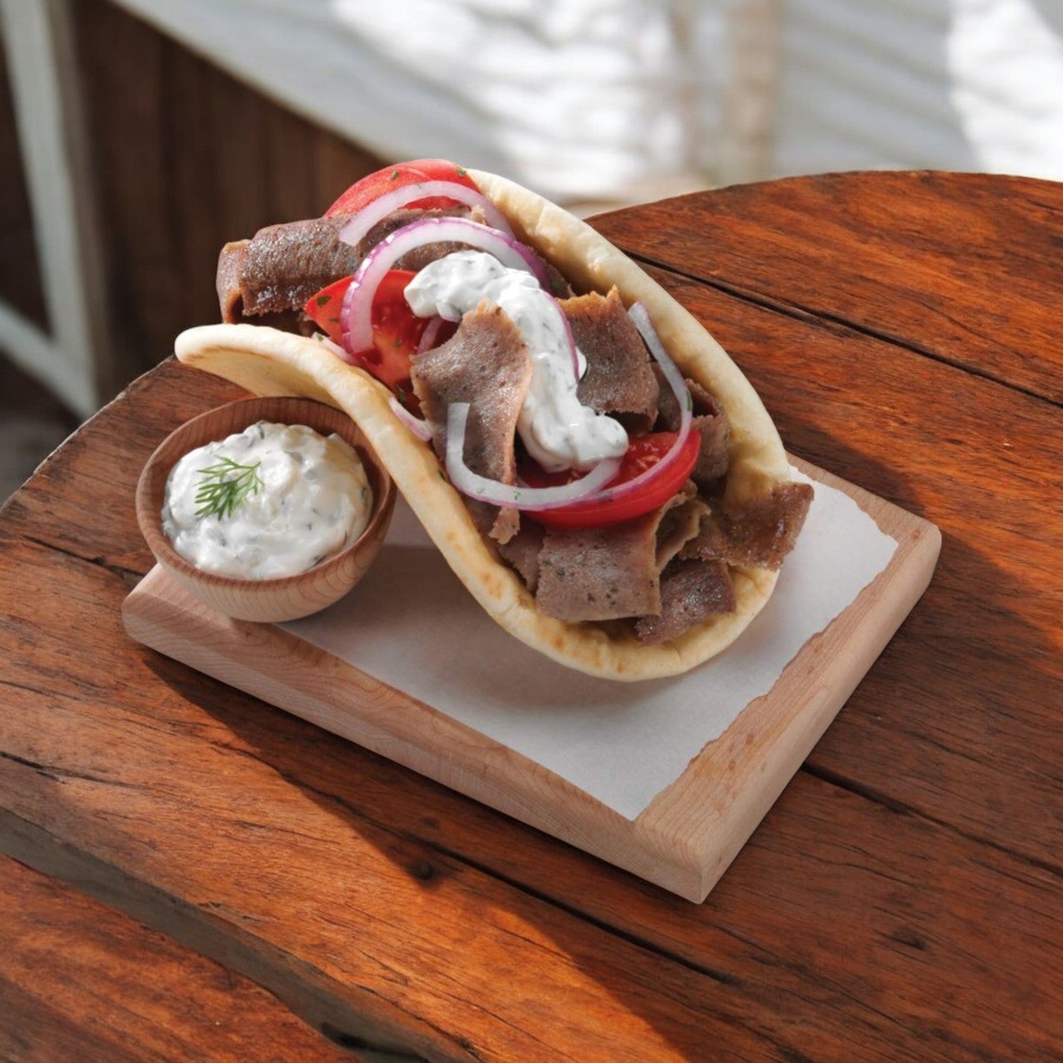 A gyro featuring Devanco's Halal Gyro Slices, onions, tomatoes, and tzatziki sauce with Mediterranean spices on pita bread, served on a rustic wooden board with extra tzatziki.