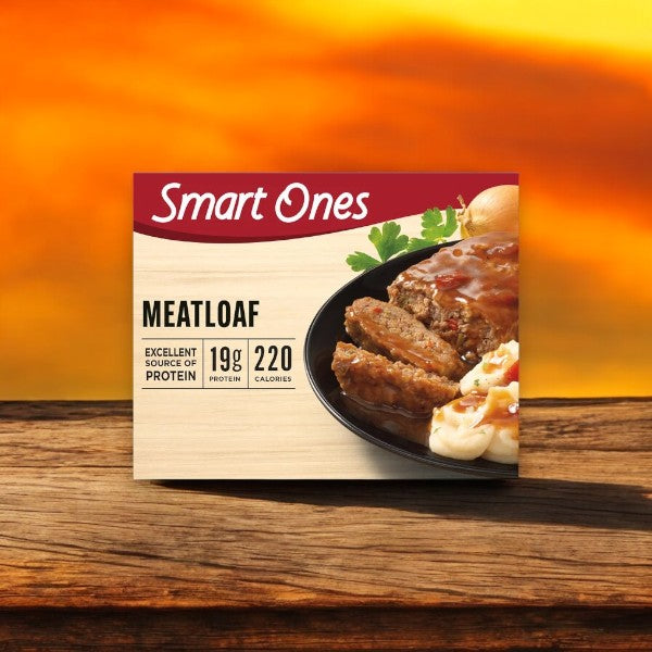A box of Smart Ones Chicken Parmesan with Spaghetti rests on a wooden surface, featuring a frozen meal with marinara sauce and mozzarella cheese. It's the epitome of microwaveable convenience. A blurred sunset in the background adds warmth to this culinary delight.