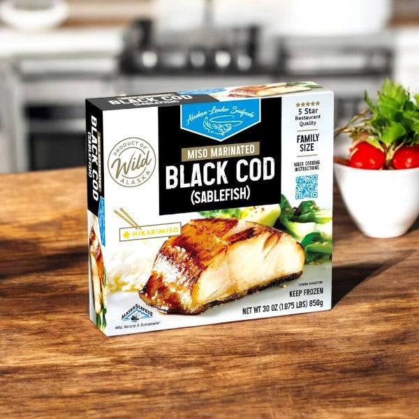 A box of Alaskan Leader Seafoods Black Cod, Miso Marinated (30 oz, 1 Pack) sits on a wooden table with tomatoes and greens in the background.