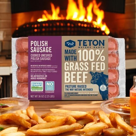 The Teton Waters Ranch Polish Sausage, made with 100% grass-fed beef and premium ingredients, is displayed before a cozy fireplace alongside a plate of fries.