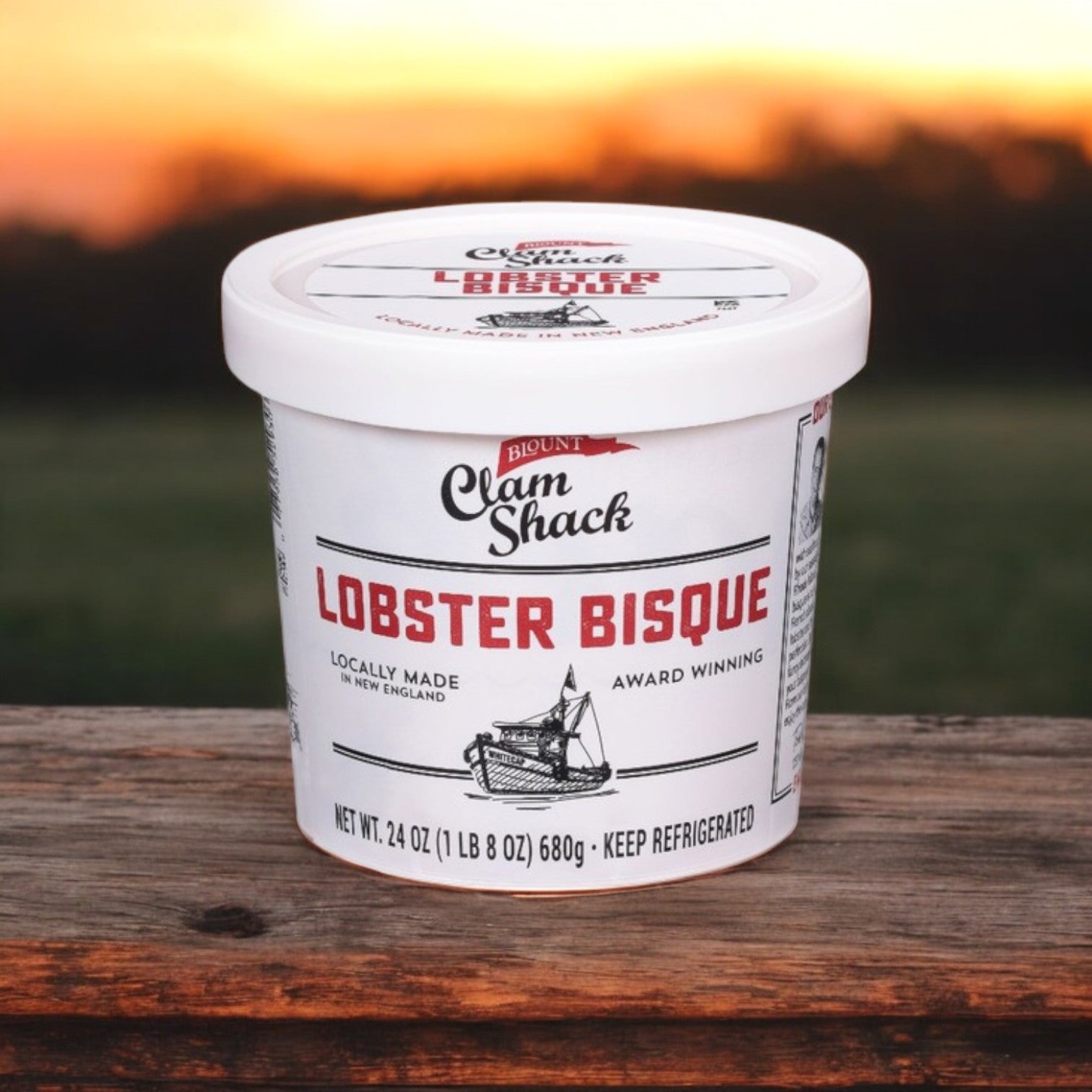 A 24 oz. container of Blount Fine Foods' Blount Clam Shack Lobster Bisque Soup with premium lobster is placed on a wooden surface against a sunset backdrop.