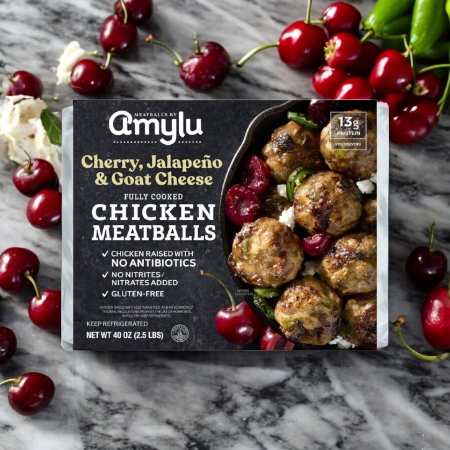 A 40 oz pack of Amylu Cherry, Jalapeno, and Goat Cheese Chicken Meatballs sits on a marble surface surrounded by fresh cherries and jalapeños.