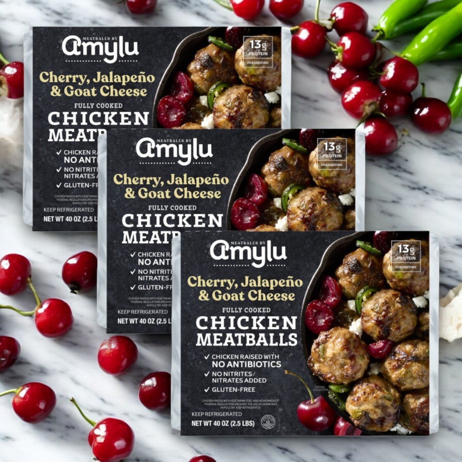 Three packs of Amylu Cherry, Jalapeno, and Goat Cheese Chicken Meatballs (40 oz) are surrounded by vibrant cherries and peppers on a gray marble surface.
