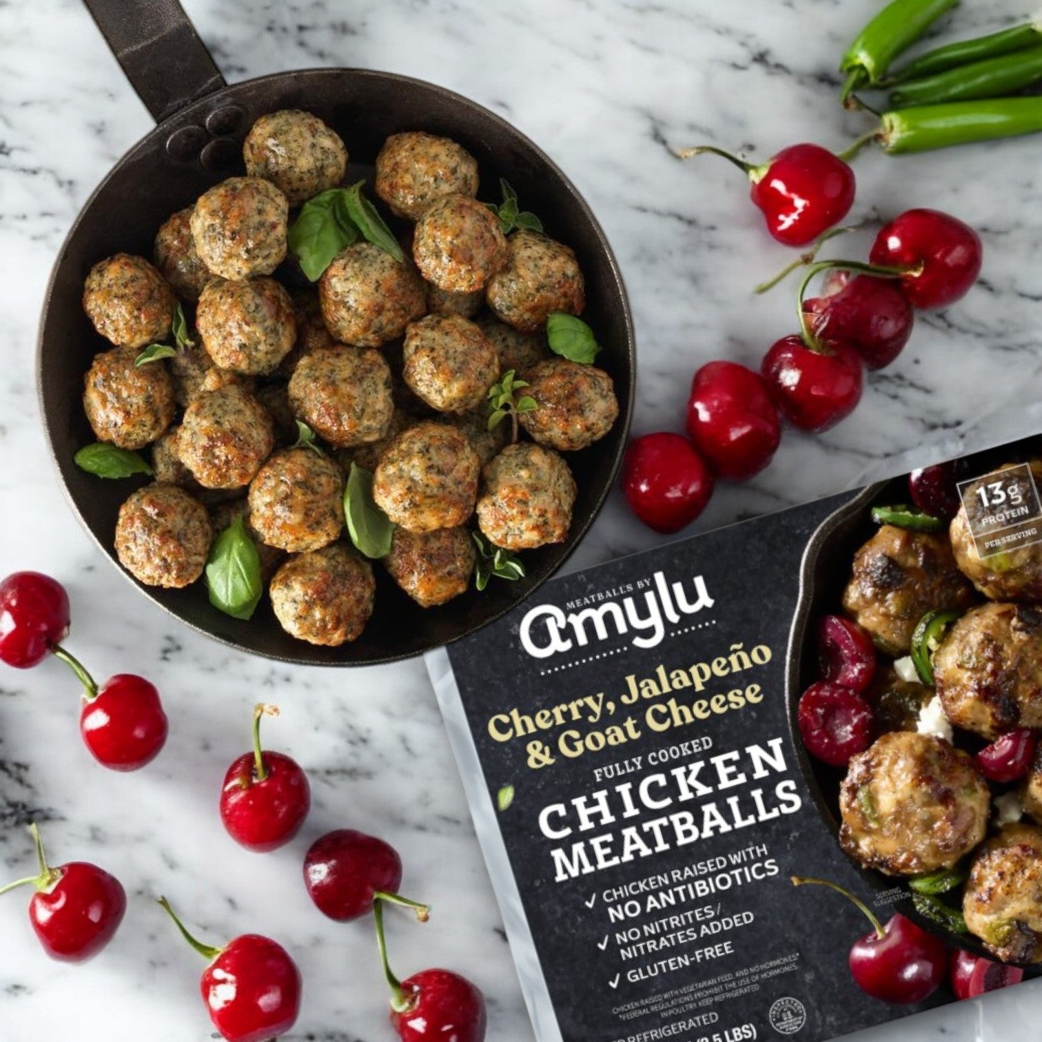 A sizzling pan of chicken meatballs with herbs sits beside a package of Amylu Cherry, Jalapeño, and Goat Cheese Chicken Meatballs (40 oz, 3 Packs), artfully surrounded by vibrant cherries and fresh green chilies on a sleek marble surface.