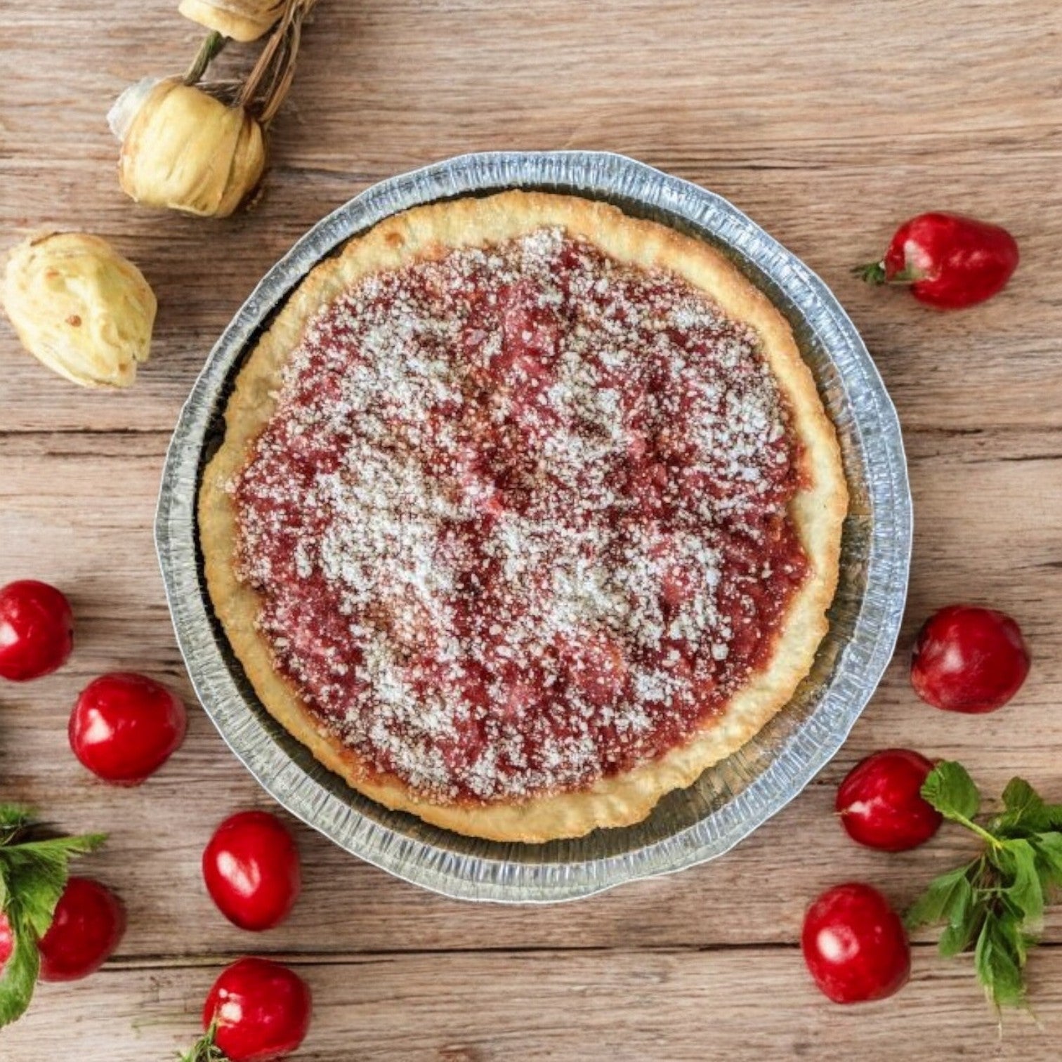 A Lou Malnati's Deep Dish Pizza Sausage, 27.5 oz., in a foil tray with red tomato sauce, grated cheese, and Italian sausage, is placed on a wooden surface adorned with fresh tomatoes and buttery flaky crust pastry puffs.