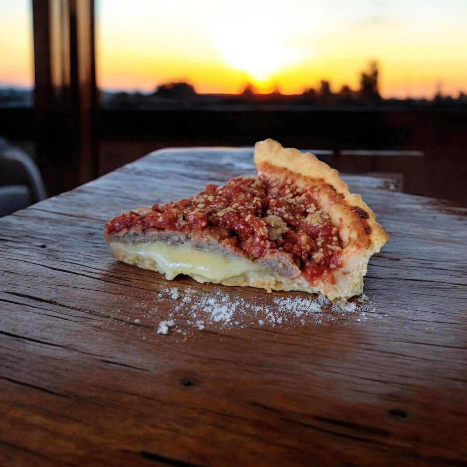 A Lou Malnati's Sausage Deep Dish Pizza slice, adorned with tomato sauce and melty cheese, sits on a wooden surface, showcasing its buttery flaky crust as a sunset casts warm hues in the background.