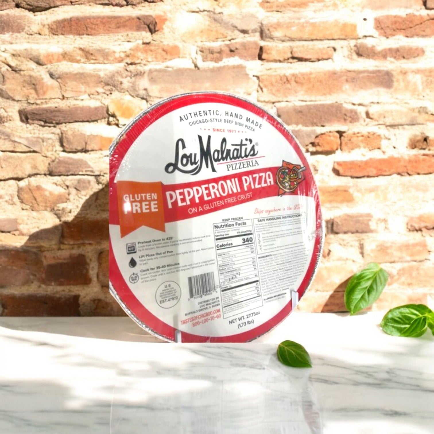 The Lou Malnati's Deep Dish Gluten Free Pack! - 2 Pizzas! is showcased against a brick wall, garnished with fresh basil leaves on a white surface, embodying the authentic flavor of Chicago Deep Dish in every bite.
