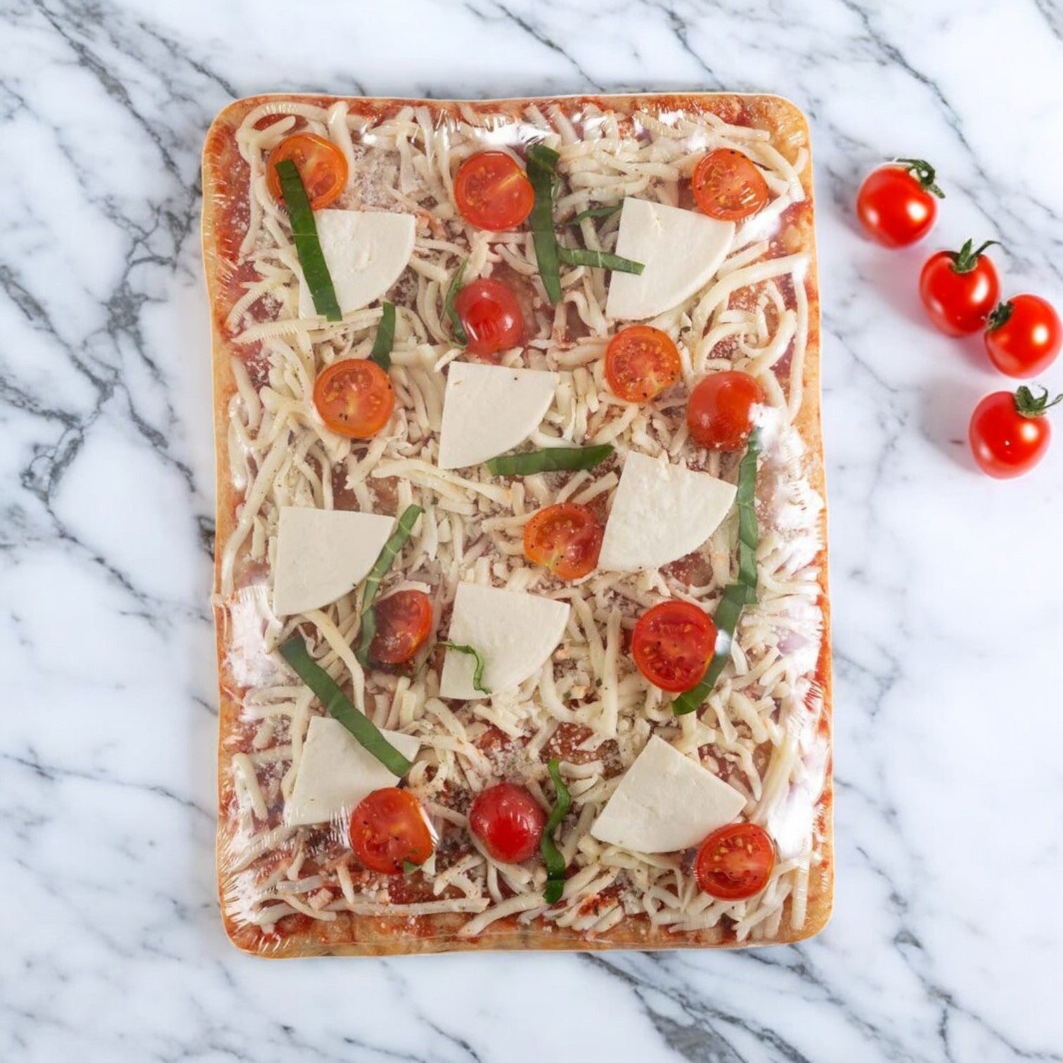A rectangular Massa Pizza Co. Margherita Bakery Pizza, featuring handcrafted dough topped with cheese, tomato slices, and basil leaves, is displayed on a marble surface next to three whole cherry tomatoes.
