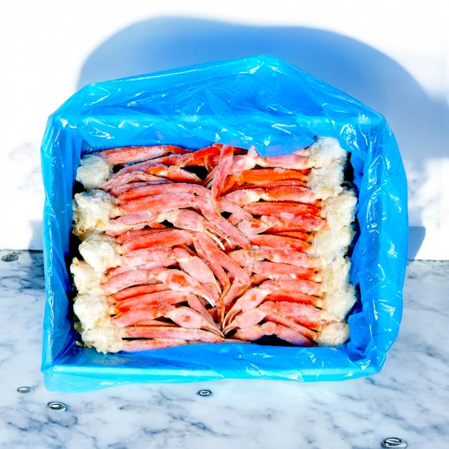 The Olivia Wild Caught Snow Crab Clusters from IFC Seafood, fully cooked and weighing 10 lbs, sit ready to enjoy in a blue plastic-lined box on a marble surface, highlighting their premium appeal.