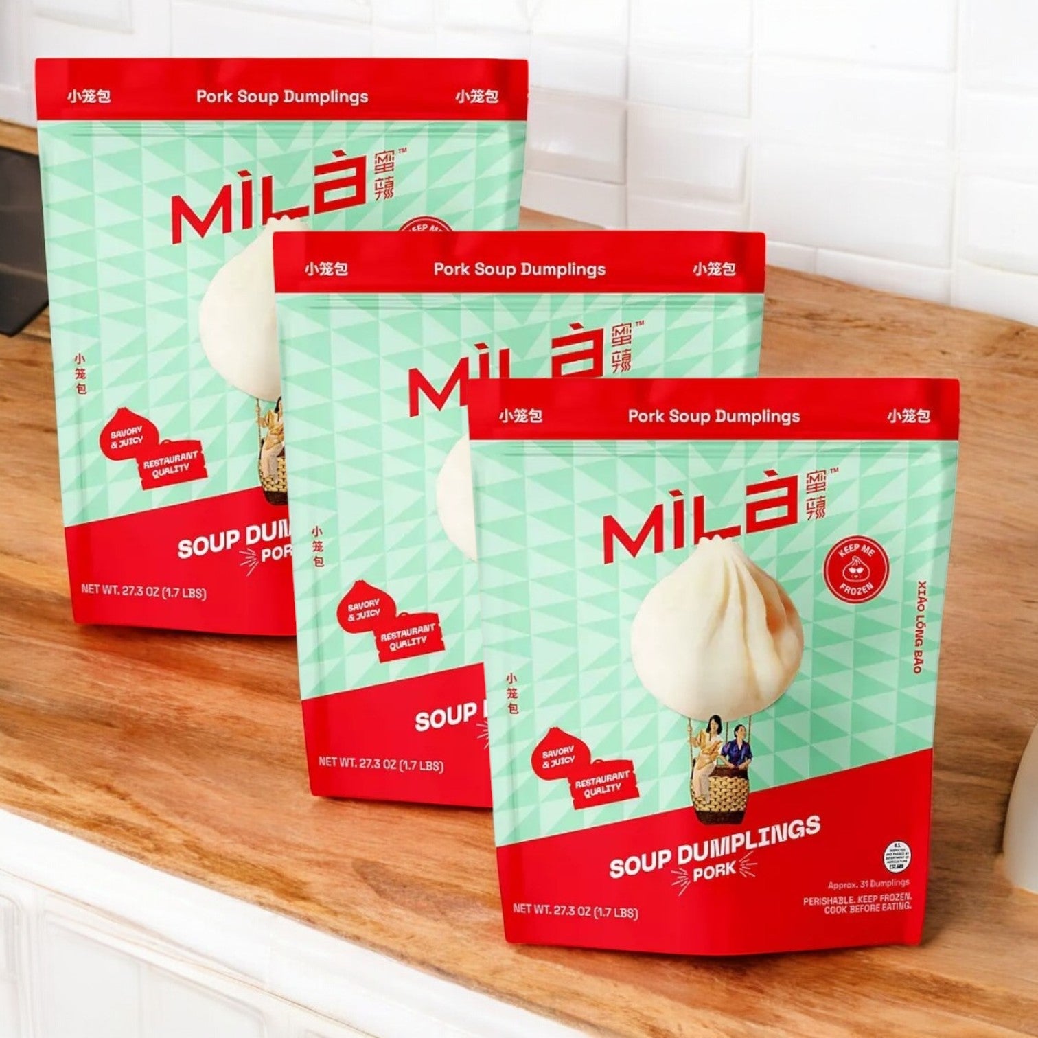 Three Mila Pork Soup Dumplings packages, each with 31 frozen dumplings, sit invitingly on a wooden countertop in the white-tiled kitchen, offering a delicious xiao long bao experience. Perfect for quick meals, they promise authentic flavor with every bite.
