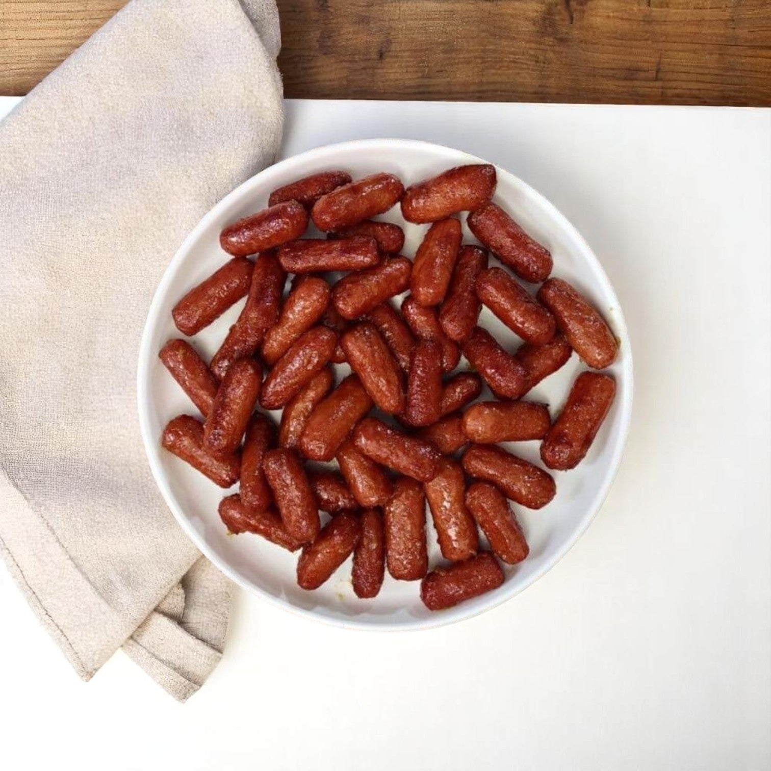 A white bowl filled with Nathan’s Famous Cocktail Franks – 1200 Count, 1 Case sits on a white surface beside a folded beige napkin and a wooden tabletop.