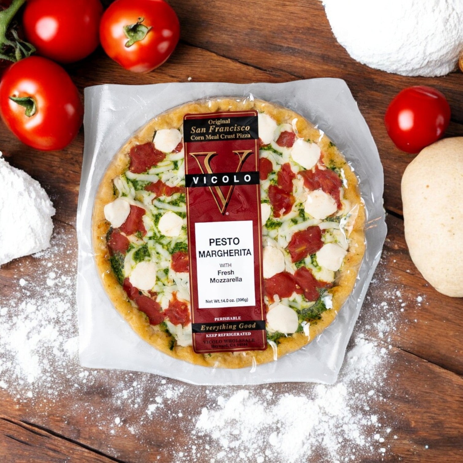 A Vicolo Pesto Margherita with Fresh Mozzarella pizza rests on a wooden surface, surrounded by tomatoes, flour, and dough. It features basil pesto and fresh mozzarella for a delightful taste experience.
