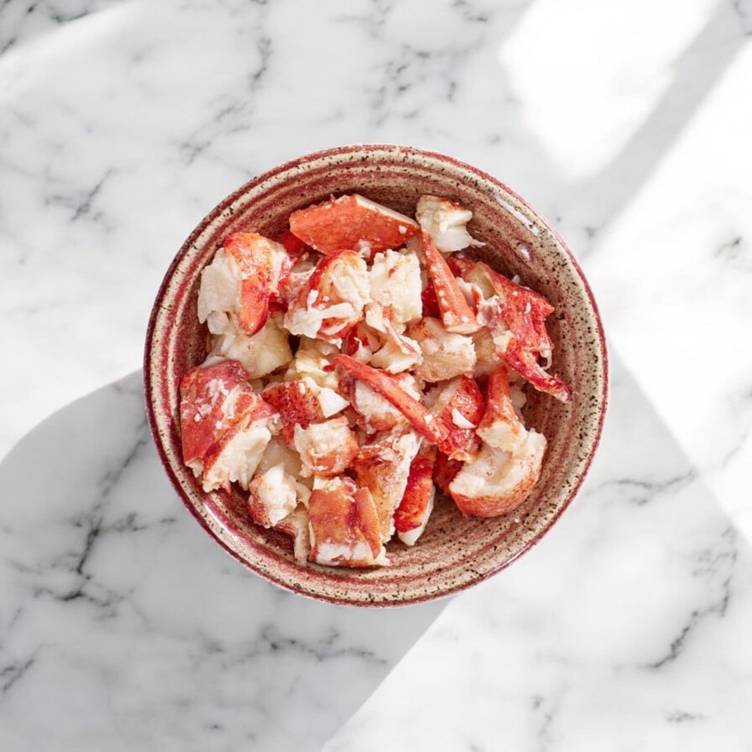 A bowl of Easy Lunches Maine Lobster Meat Claw and Knuckle - 2 Pounds rests on a marble surface, with natural light casting captivating shadows.