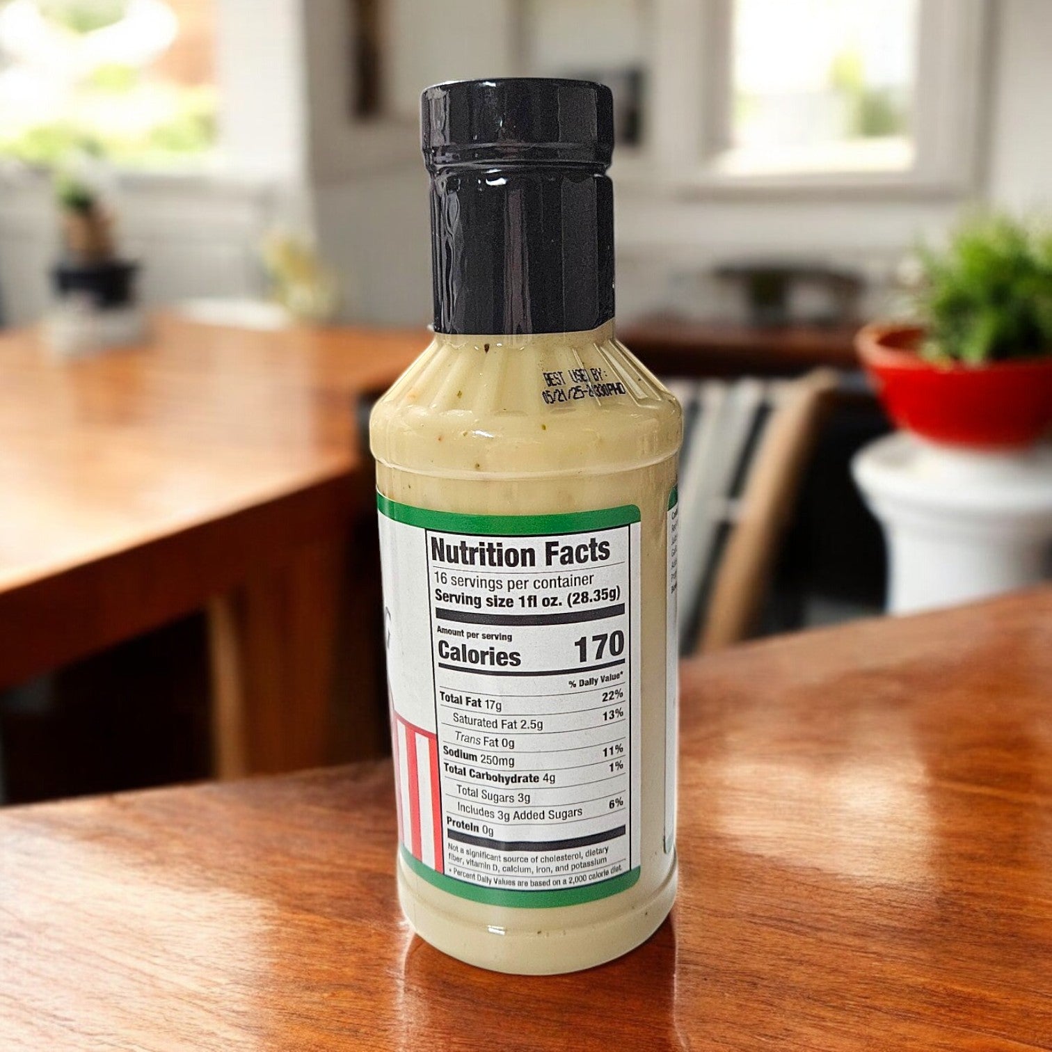 A 16 oz. bottle of Portillo's House Dressing stands on a wooden table, displaying its nutrition facts label. The background, a blurred indoor scene with scattered furniture, evokes the cozy charm of Chicago-style cuisine.
