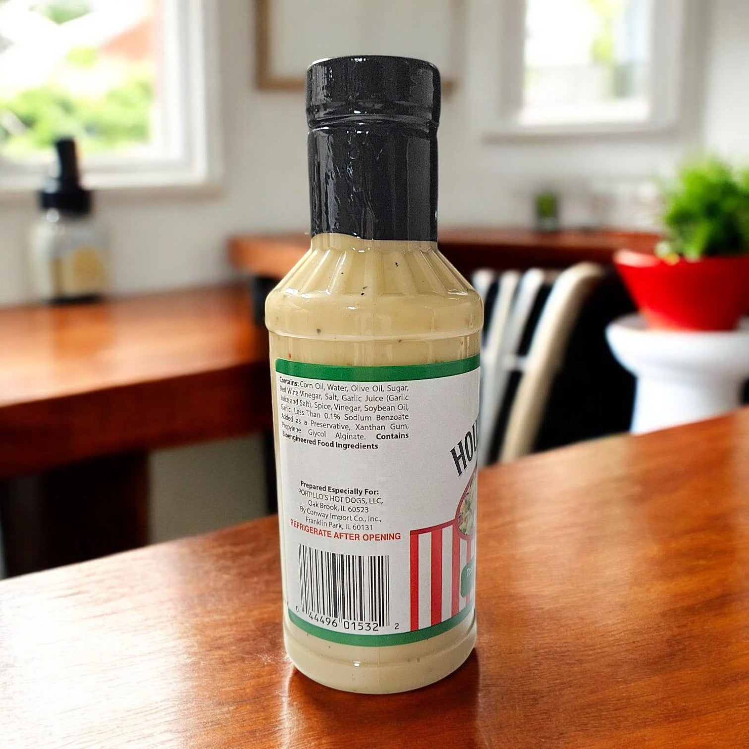 A 16 oz. bottle of Portillo's House Dressing sits on a wooden table, basking in the glow from bright windows beside a lush plant.