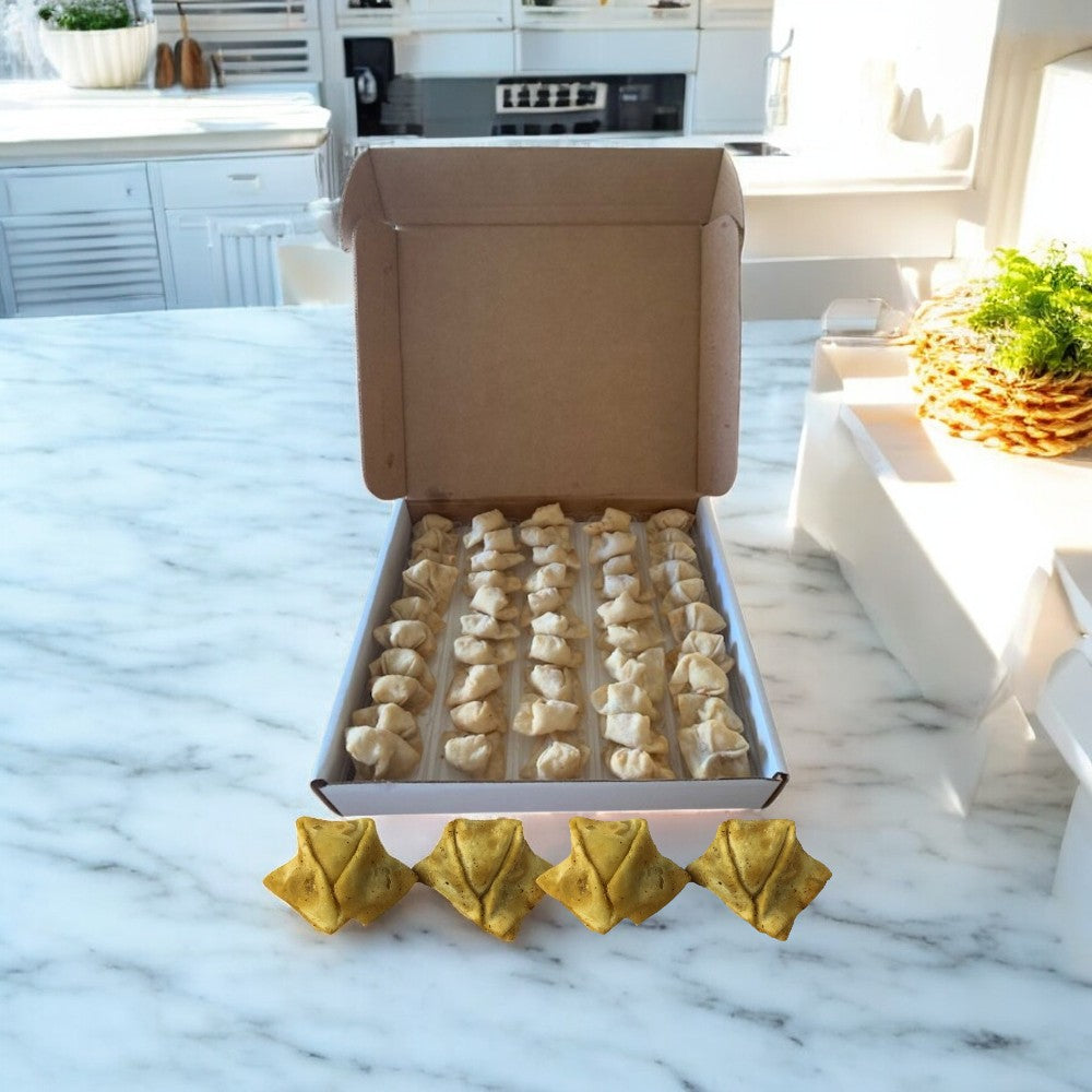An open cardboard box of Sysco Crab Rangoon-Sysco Appetizer, with each piece crafted in a delicate wonton wrapper, sits on a marble countertop. Four yellow star-shaped items are placed in front, with kitchen appliances in the background completing this enticing frozen appetizer display.