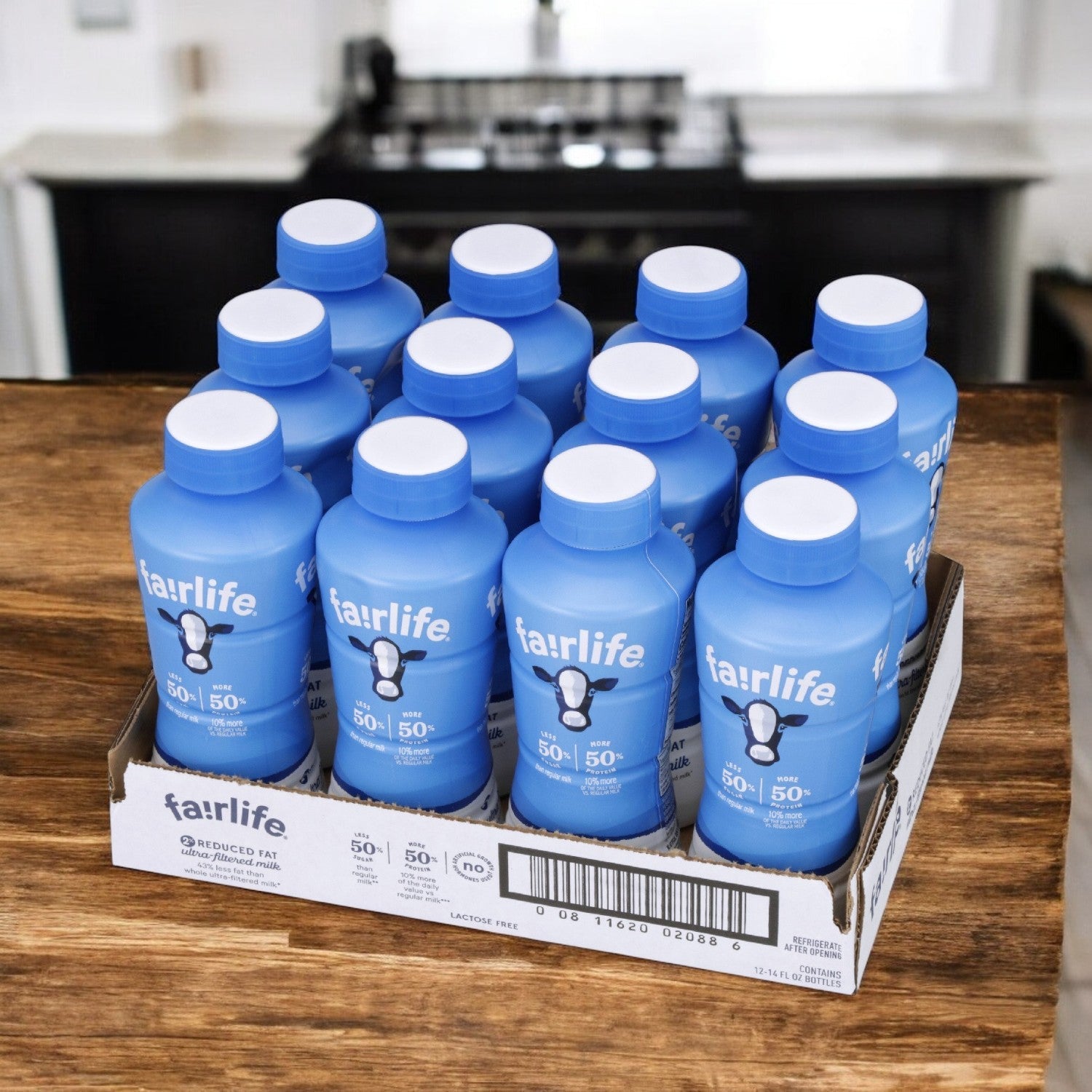 A cardboard tray with twelve blue bottles of Fairlife 2% Ultra-Filtered Reduced Fat Milk, each with a white cap, sits on a wooden surface in a kitchen. The lactose-free milk by FairLife is high in protein, ideal for those seeking delicious nutrition.