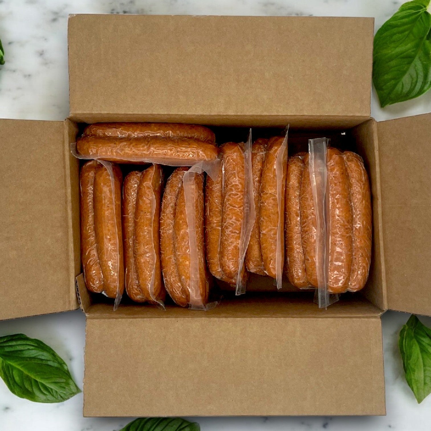 An open cardboard box of Amylu Chicken Sausage Links – Apple & Maple, 72 Count (10 lbs.), sits on a marble surface with green basil leaves around it, exuding a delicious apple and maple aroma.