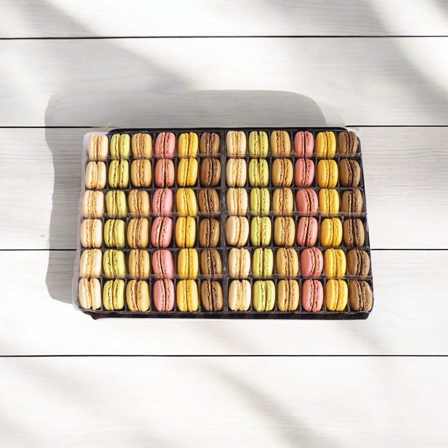A rectangular Pasquier box with rows of colorful Cookie Macaron Classic French, a luxurious dessert featuring 144 delightful macarons, is elegantly displayed on a white wooden surface.