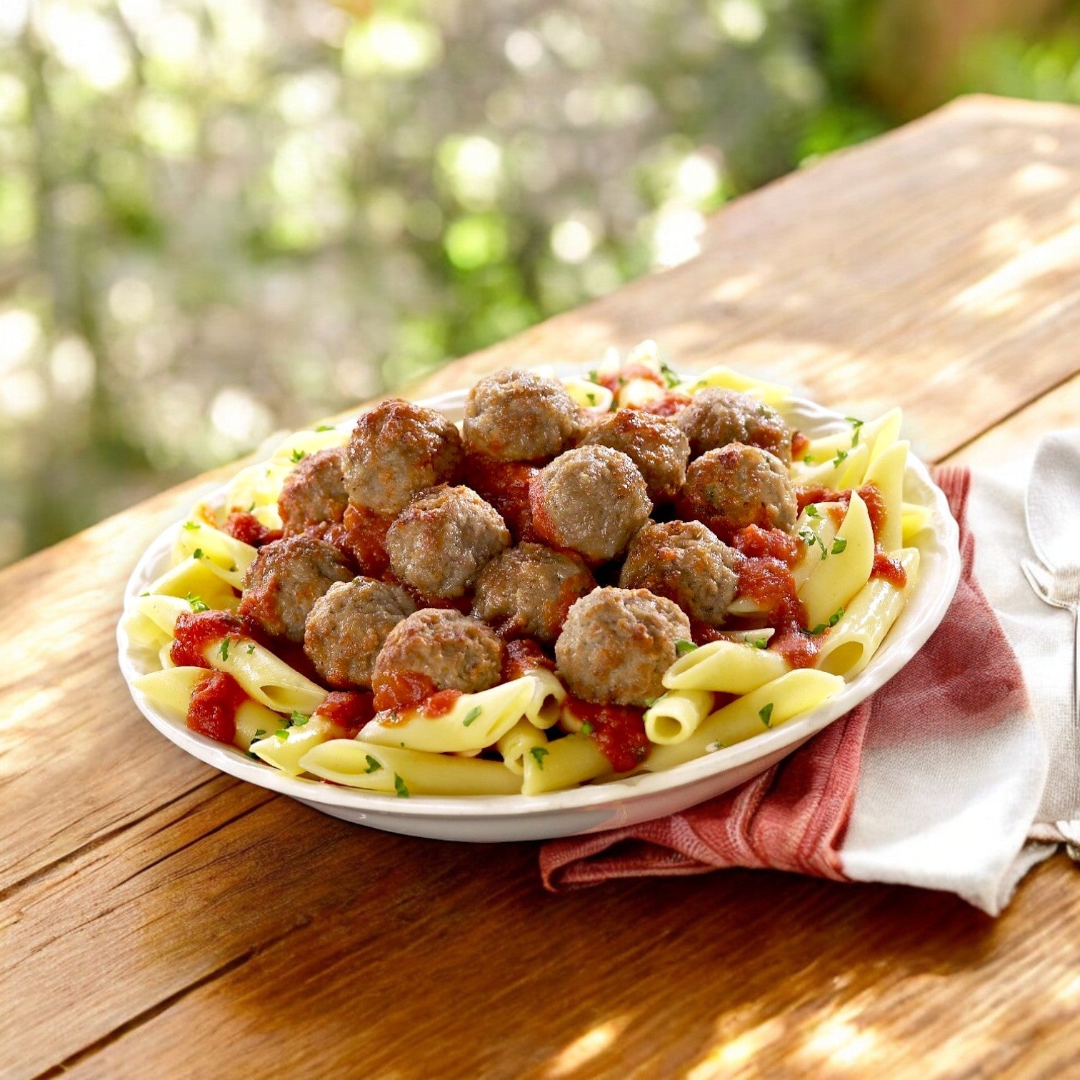 A plate of penne pasta topped with tomato sauce and Rosina Halal Beef & Chicken Meatballs sits on a wooden table beside a folded napkin, offering an authentic Italian flavor.