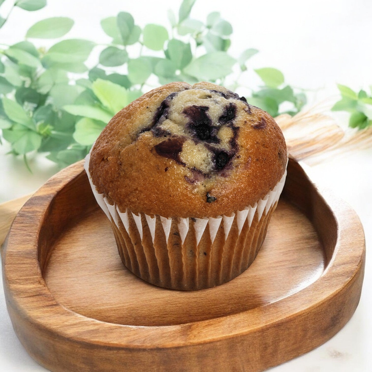 An Otis Spunkmeyer Muffin Blueberry Wild from a case of 96 sits on a wooden tray amid green foliage—the perfect breakfast treat to start your day with free shipping!.
