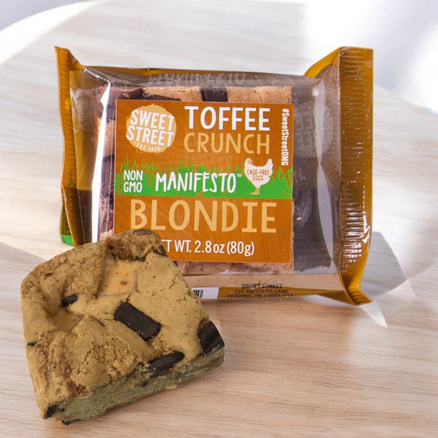 A package of Sweet Street Blondie Toffee Crunch 2.8 oz. Brownies, 48 count, rests on a wooden surface with an unwrapped piece showcasing rich chocolate chunks—a delightful addition to premium dessert selections.
